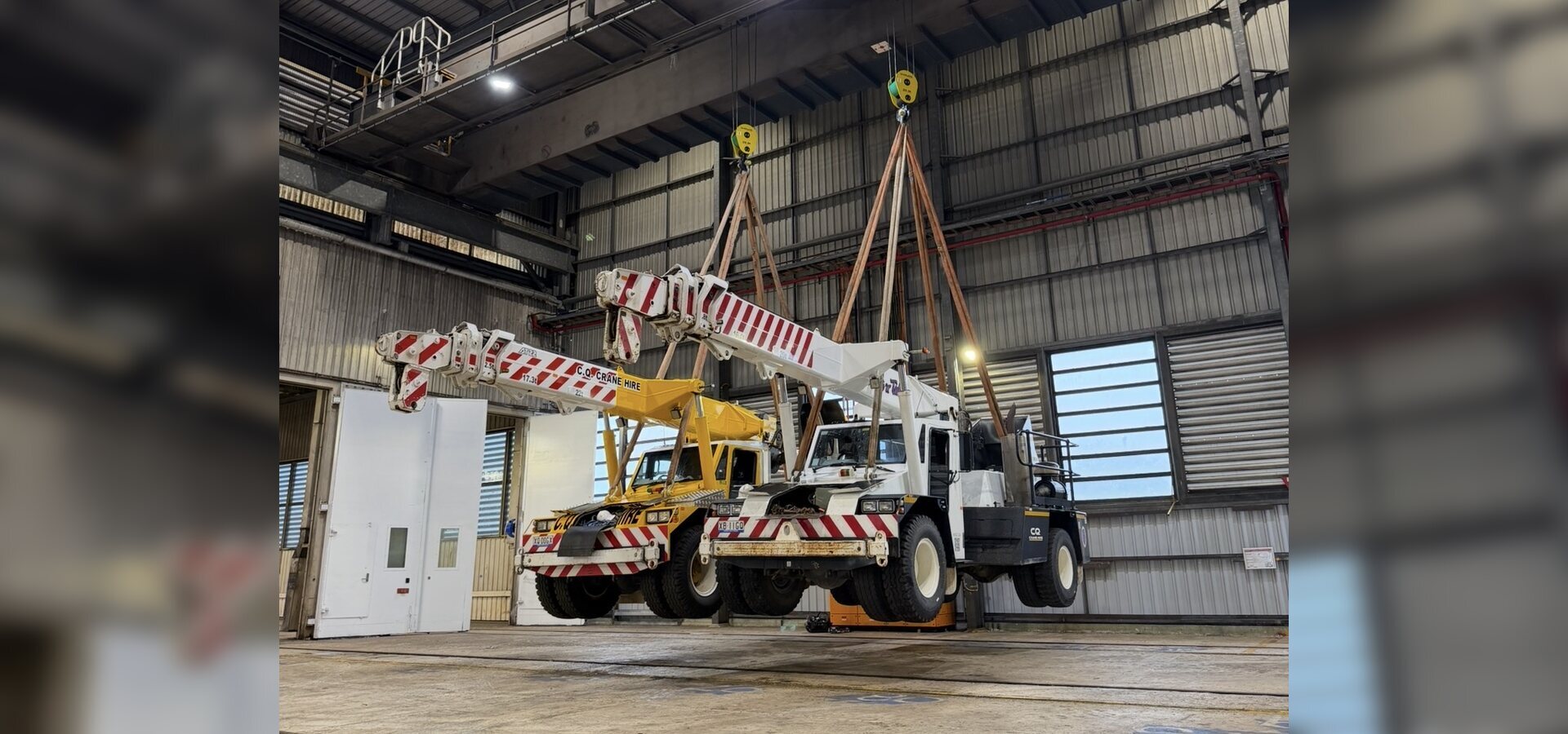 Overhead crane lifting two mobile cranes inside an industrial workshop